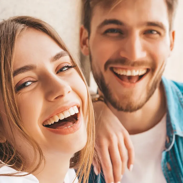 Un noi i una noia joves somriuen de manera natural i feliç, mostrant dents cuidades i sanes. Representen confiança i benestar després d’un tractament dental.